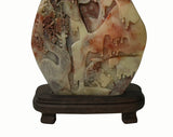 A carved stone sculpture depicting a landscape with trees and a figure, displayed on a dark wooden stand against a plain white background.