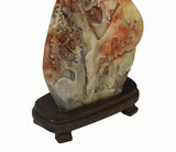 A carved decorative stone sculpture with a landscape motif, displayed on a dark wooden stand against a plain white background.