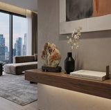 A modern living room interior with a floating wooden console table, a black vase with dried branches, a decorative stone sculpture on a wooden base, and a book. A gray sectional sofa, large window view of a city skyline, and textured beige walls are visible.